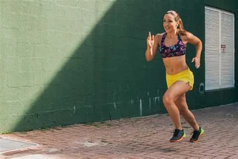 Kobieta w stroju sportowym biegnie chodnikiem wzdłuż ciemnozielonej ściany w słoneczny dzień.