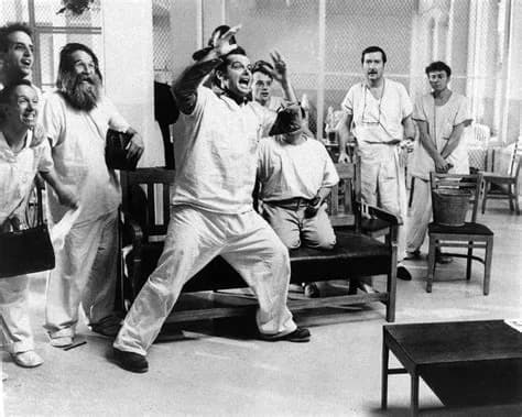 Czarno-biały kadr z filmu „Lot nad kukułczym gniazdem” (1975): pacjenci oddziału psychiatrycznego w szpitalnych ubraniach, jeden z nich unosi zaciśnięte pięści w emocjonalnym geście.